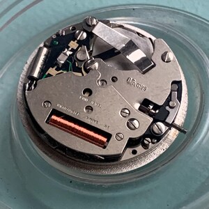 Op de afbeelding: Gedetailleerd beeld van een uurwerk, met zijn complexe interne werking. Het zilveren mechanisme bevat een koperen spoel, talrijke schroeven en de tekst "UNADJUSTED SWISS". Het uurwerk is gehuisvest in een heldere, cirkelvormige container.