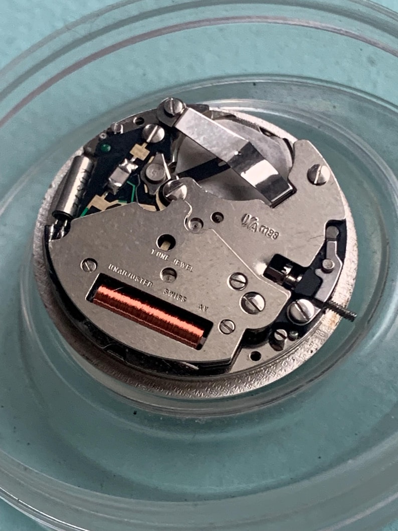 Op de afbeelding: Gedetailleerd beeld van een uurwerk, met zijn complexe interne werking. Het zilveren mechanisme bevat een koperen spoel, talrijke schroeven en de tekst "UNADJUSTED SWISS". Het uurwerk is gehuisvest in een heldere, cirkelvormige container.