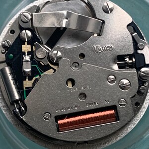 Op de afbeelding: Close-up van een uurwerk met zilverkleurige metalen componenten. De tekst "VA m99", "1 ONE JEWEL", "UNADJUSTED SWISS AV" is zichtbaar. Er is ook een koperkleurige spoel aanwezig.