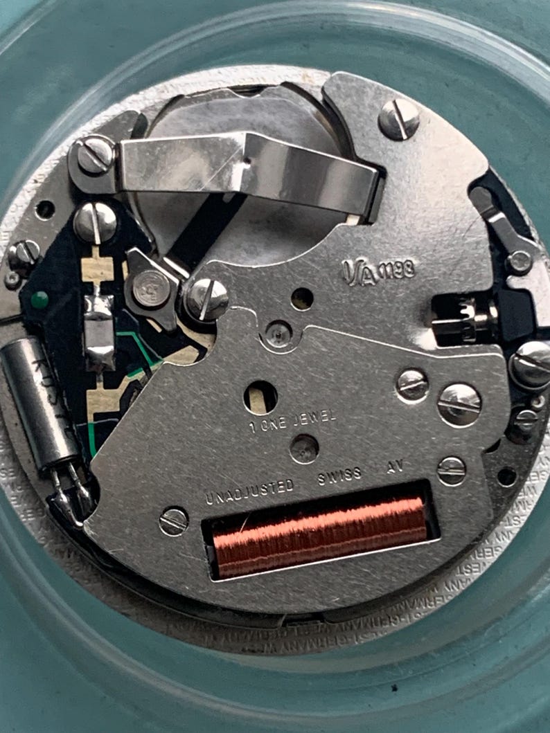 Op de afbeelding: Close-up van een uurwerk met zilverkleurige metalen componenten. De tekst "VA m99", "1 ONE JEWEL", "UNADJUSTED SWISS AV" is zichtbaar. Er is ook een koperkleurige spoel aanwezig.