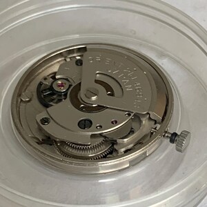 May include: A close-up of a watch movement, featuring intricate gears, a rotor, and small jewels. The movement is encased in a clear plastic container. The text "ORIENT 21 JEWELS JAPAN" is visible on the rotor.