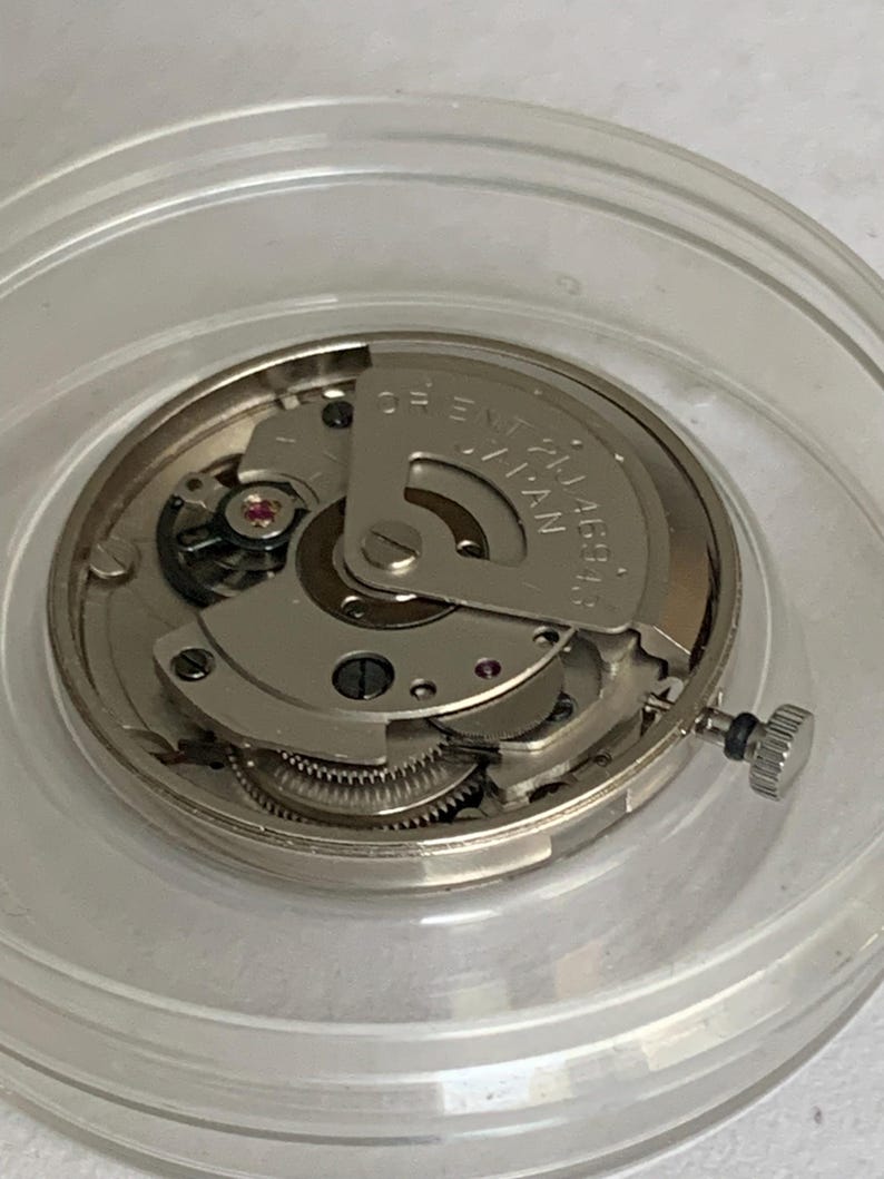 May include: A close-up of a watch movement, featuring intricate gears, a rotor, and small jewels. The movement is encased in a clear plastic container. The text "ORIENT 21 JEWELS JAPAN" is visible on the rotor.
