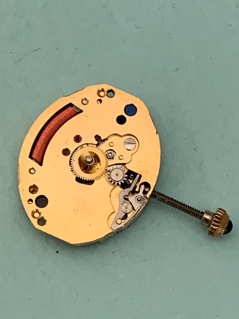 May include: Close-up of a gold-colored watch movement with intricate gears, a red coil, and a screw with a black head. The watch mechanism is set against a light blue background.