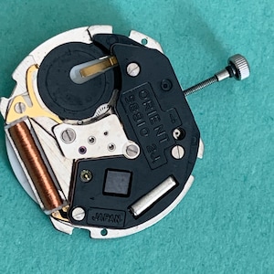 May include: A close-up of a watch movement with the text "ORIENT 58910 2J" and "JAPAN" visible. The movement features a black and silver design with gold and copper-coloured components. A small screw is attached to the side.