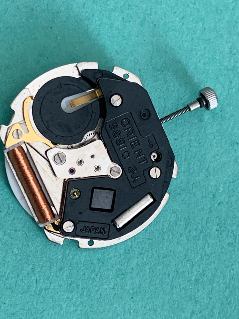 May include: A close-up of a watch movement with the text "ORIENT 58910 2J" and "JAPAN" visible. The movement features a black and silver design with gold and copper-coloured components. A small screw is attached to the side.