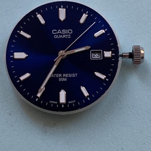 May include: A round Casio quartz watch dial with a deep blue sunburst finish, silver-tone hour markers, and silver hands. The dial features a date window at the three o'clock position, the text CASIO QUARTZ, WATER RESIST 50M, and JAPAN MOVT, with a metal crown.