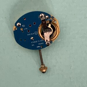 May include: A close-up of a watch movement with a blue circuit board. The movement has gold and silver components, including a battery holder and a screw. The text "SWISS SIX 6 JEWELS" is visible.
