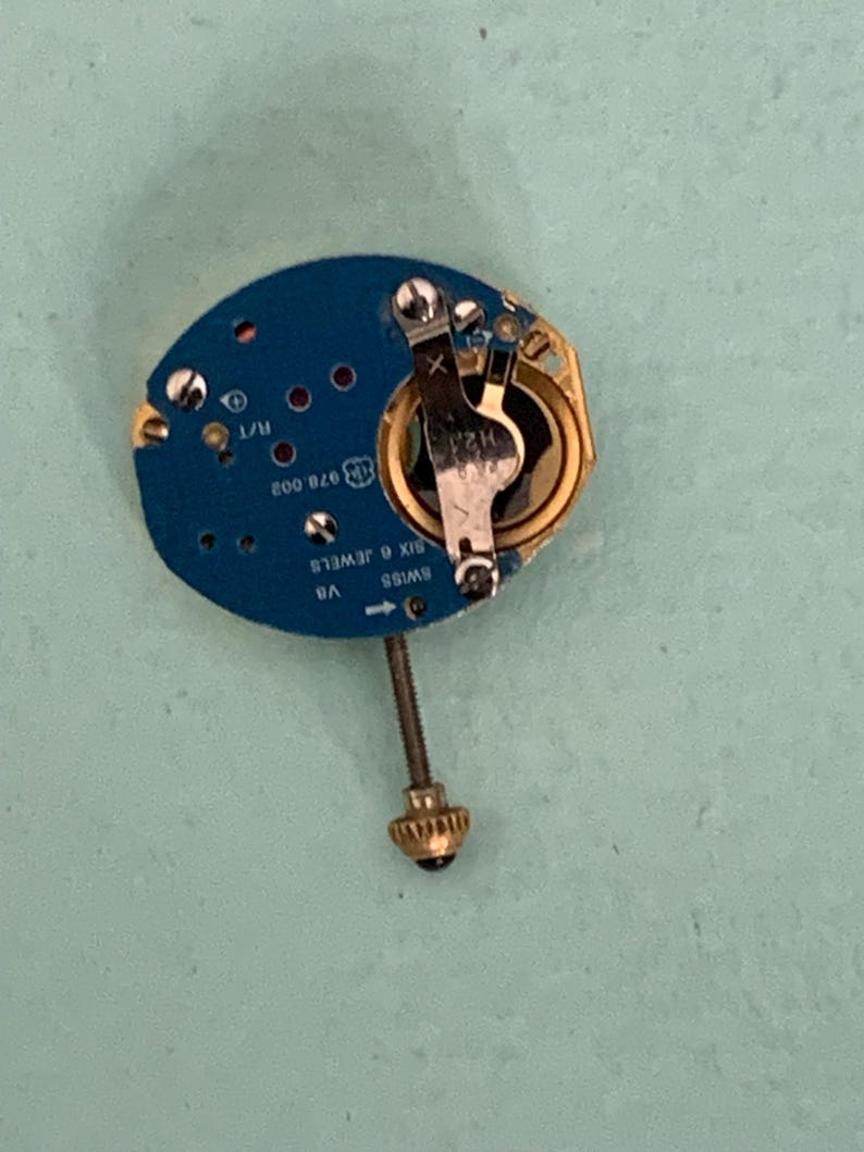 May include: A close-up of a watch movement with a blue circuit board. The movement has gold and silver components, including a battery holder and a screw. The text "SWISS SIX 6 JEWELS" is visible.