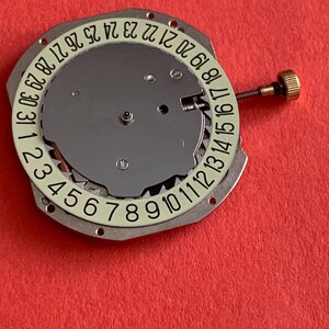 K&ouml;nnte beinhalten: Eine Uhrwerkkomponente mit einer silberfarbenen zentralen Scheibe und einem wei&szlig;en Ring mit schwarzen Zahlen von 1 bis 31. Eine kleine goldfarbene Schraube ist angebracht. Die Komponente befindet sich vor einem roten Hintergrund.
