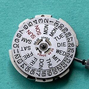 May include: A close-up of a watch movement with a white dial featuring the days of the week and numbers 1-31. The dial has a silver metal frame and a small crown. The text "334EG" and "52-45" are visible.