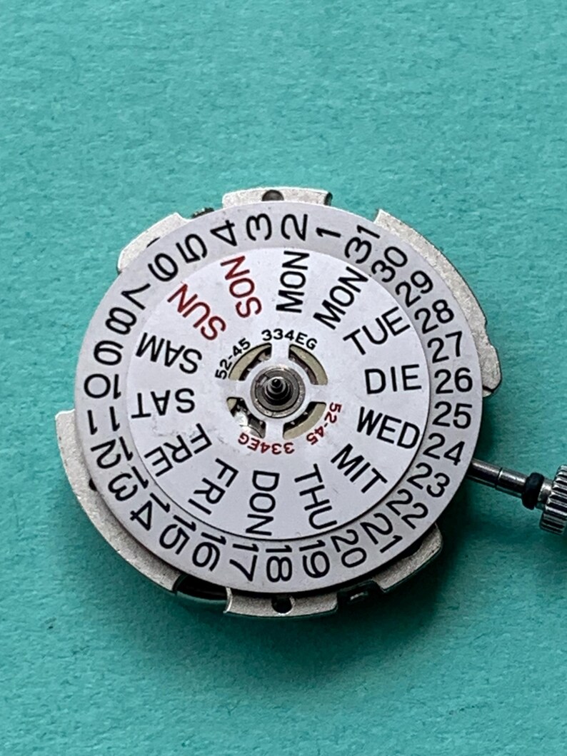 May include: A close-up of a watch movement with a white dial featuring the days of the week and numbers 1-31. The dial has a silver metal frame and a small crown. The text "334EG" and "52-45" are visible.