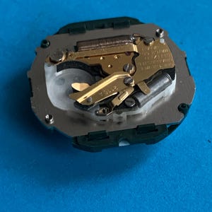 May include: A detailed view of a watch movement, featuring gold-toned gears and a silver base, set against a blue backdrop. The intricate mechanism showcases fine details, typical of precision watchmaking.