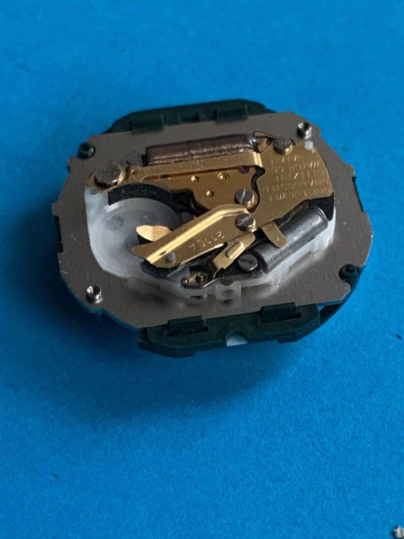 May include: A detailed view of a watch movement, featuring gold-toned gears and a silver base, set against a blue backdrop. The intricate mechanism showcases fine details, typical of precision watchmaking.