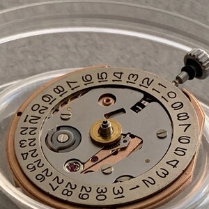 Puede incluir: Primer plano de un movimiento de reloj con esfera plateada y anillo dorado. La esfera muestra n&uacute;meros negros que indican los d&iacute;as del mes. El mecanismo incluye peque&ntilde;os engranajes, una joya y una tija de cuerda.