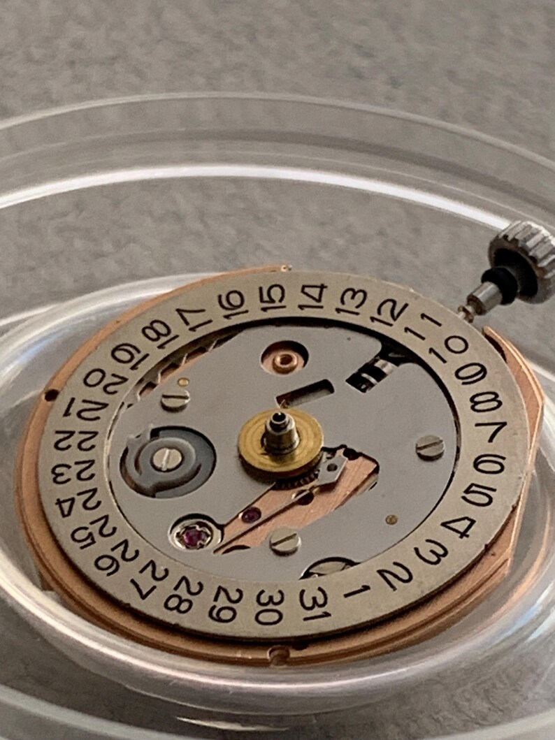 Puede incluir: Primer plano de un movimiento de reloj con esfera plateada y anillo dorado. La esfera muestra n&uacute;meros negros que indican los d&iacute;as del mes. El mecanismo incluye peque&ntilde;os engranajes, una joya y una tija de cuerda.