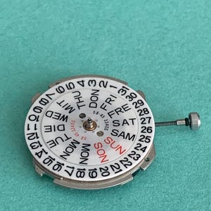 May include: A watch movement with a white dial featuring black numerals and day indicators in English and German. The day of the week is indicated in red. A small metal screw extends from the side.