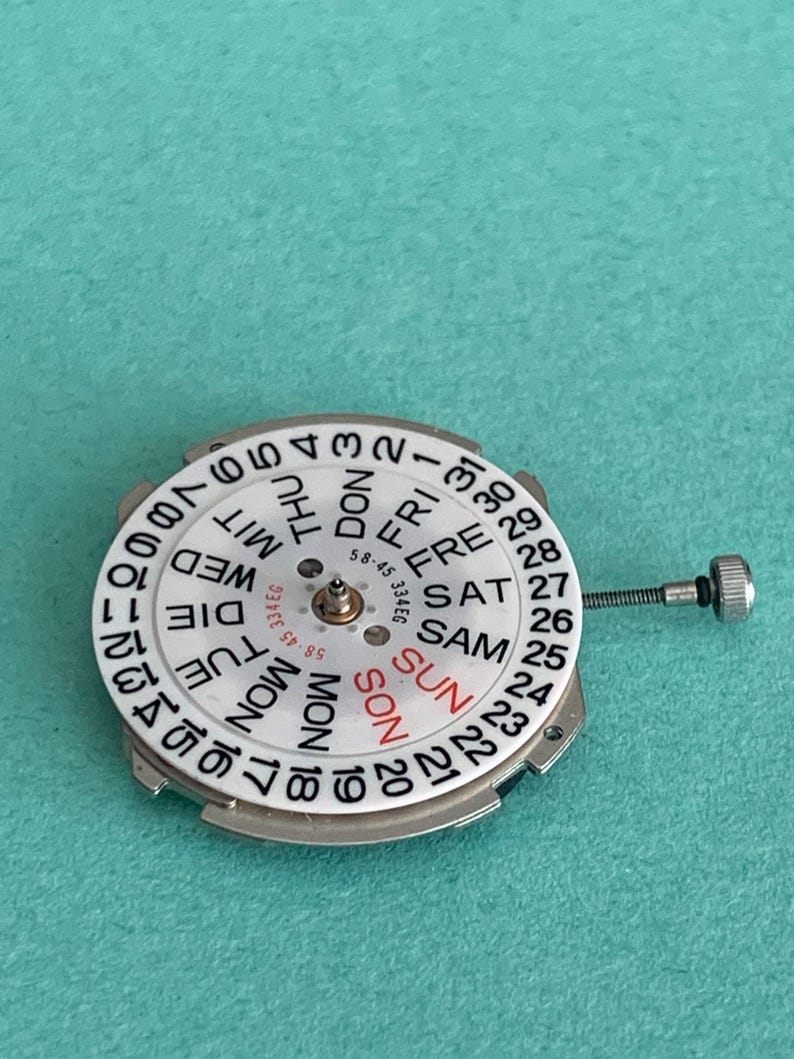 May include: A watch movement with a white dial featuring black numerals and day indicators in English and German. The day of the week is indicated in red. A small metal screw extends from the side.