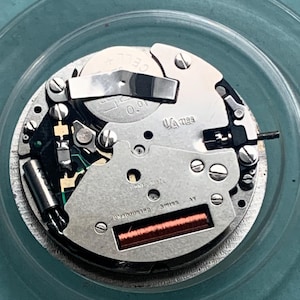Op de afbeelding: Close-up van een uurwerk met zilveren en gouden componenten. Het mechanisme bevat een batterij, een spoel en de tekst "UNADJUSTED SWISS". Het uurwerk staat tegen een turquoise achtergrond.