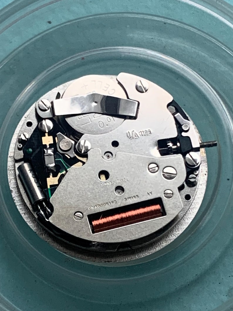 Op de afbeelding: Close-up van een uurwerk met zilveren en gouden componenten. Het mechanisme bevat een batterij, een spoel en de tekst "UNADJUSTED SWISS". Het uurwerk staat tegen een turquoise achtergrond.