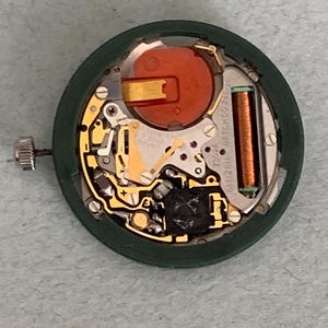 May include: A disassembled watch movement with a green outer ring. The internal components include gold-colored gears, a red battery, and a copper-colored coil. The watch crown is visible on the left side.