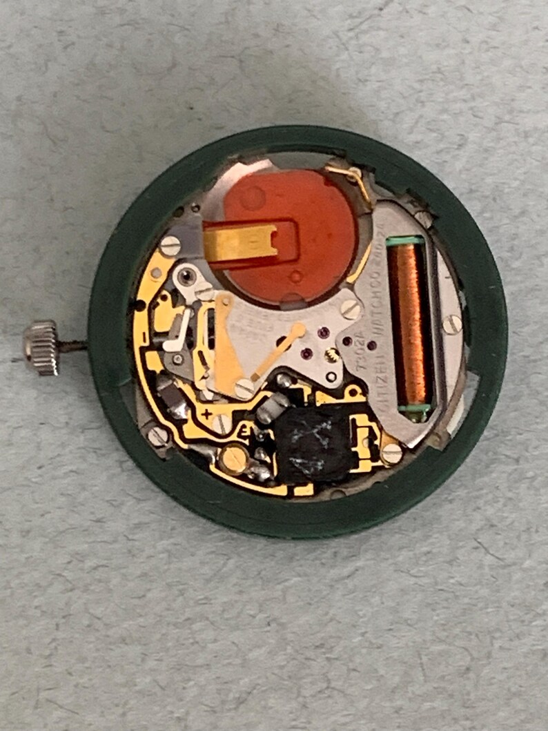 May include: A disassembled watch movement with a green outer ring. The internal components include gold-colored gears, a red battery, and a copper-colored coil. The watch crown is visible on the left side.