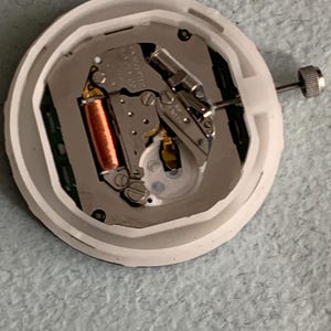 Puede incluir: Un movimiento de reloj con una carcasa de pl&aacute;stico blanco. Los componentes internos incluyen una bobina de cobre, engranajes met&aacute;licos y una esfera plateada. Una peque&ntilde;a corona plateada es visible en el lateral. Mecanismo de reloj para reparaci&oacute;n o reemplazo.