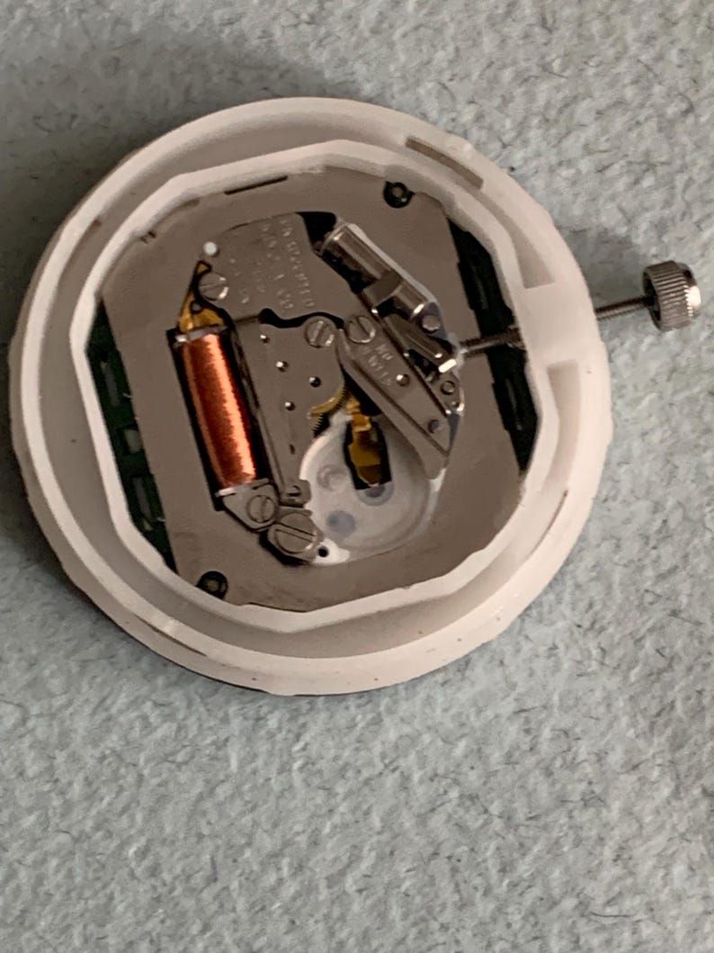 Puede incluir: Un movimiento de reloj con una carcasa de pl&aacute;stico blanco. Los componentes internos incluyen una bobina de cobre, engranajes met&aacute;licos y una esfera plateada. Una peque&ntilde;a corona plateada es visible en el lateral. Mecanismo de reloj para reparaci&oacute;n o reemplazo.