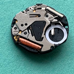 May include: Close-up of a watch movement with the text "JAPAN" and "SEIKO TIME CORP." visible. The movement features various metal components, including a copper coil and a battery compartment. The background is a teal-colored surface.