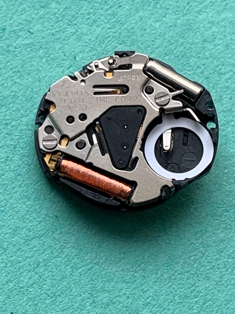 May include: Close-up of a watch movement with the text "JAPAN" and "SEIKO TIME CORP." visible. The movement features various metal components, including a copper coil and a battery compartment. The background is a teal-colored surface.