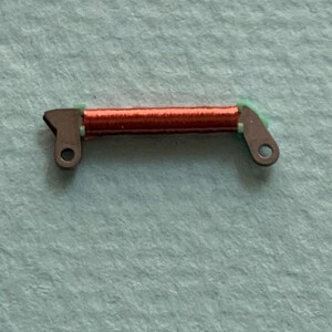 May include: A small, mechanical component featuring a coil of red wire, held between two metal brackets with green accents. The brackets have circular holes for attachment. The component is set against a light blue background.