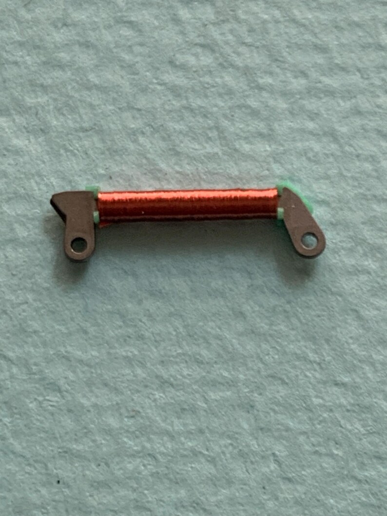May include: A small, mechanical component featuring a coil of red wire, held between two metal brackets with green accents. The brackets have circular holes for attachment. The component is set against a light blue background.