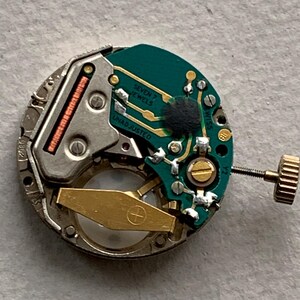 Puede incluir: Primer plano de un movimiento de reloj con una placa de circuito verde, engranajes dorados y un marco de metal plateado. El texto "SEVEN 7 JEWELS" y "UNADJUSTED" es visible. Una corona dorada est&aacute; presente.