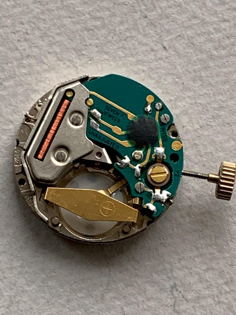 Puede incluir: Primer plano de un movimiento de reloj con una placa de circuito verde, engranajes dorados y un marco de metal plateado. El texto "SEVEN 7 JEWELS" y "UNADJUSTED" es visible. Una corona dorada est&aacute; presente.