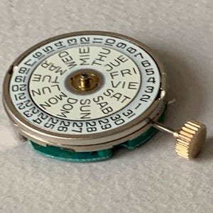 Puede incluir: Un movimiento de reloj con una esfera blanca que muestra los d&iacute;as de la semana y las fechas numeradas. La esfera tiene un centro dorado y una corona dorada. El movimiento est&aacute; sobre una base verde.