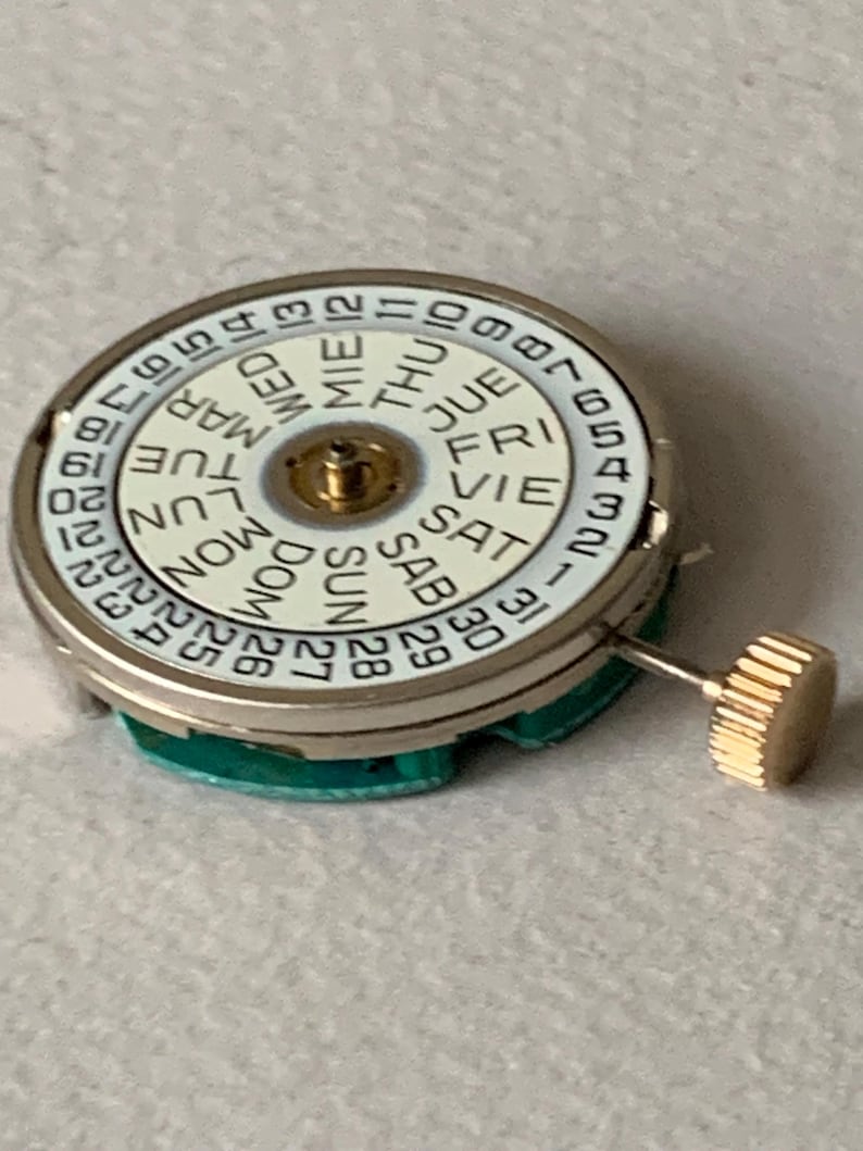 Puede incluir: Un movimiento de reloj con una esfera blanca que muestra los d&iacute;as de la semana y las fechas numeradas. La esfera tiene un centro dorado y una corona dorada. El movimiento est&aacute; sobre una base verde.