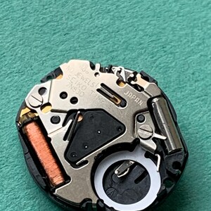 May include: A close-up of a watch movement with the text "NO JEWELS SEIKO TIME 7N83D JAPAN". The movement has silver, copper, and black components. The watch mechanism is on a green surface.