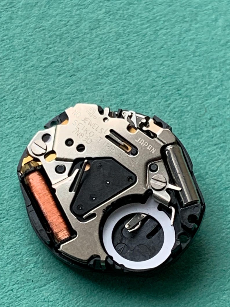 May include: A close-up of a watch movement with the text "NO JEWELS SEIKO TIME 7N83D JAPAN". The movement has silver, copper, and black components. The watch mechanism is on a green surface.