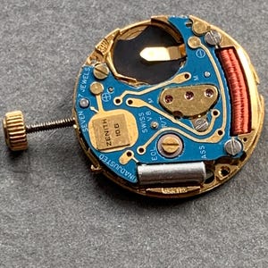 May include: Close-up of a Zenith watch movement. The watch mechanism features a gold-toned frame with a blue circuit board. The text "ZENITH 10.6" and "SWISS V8" are visible. The watch movement also has a gold-toned screw and a red coil.