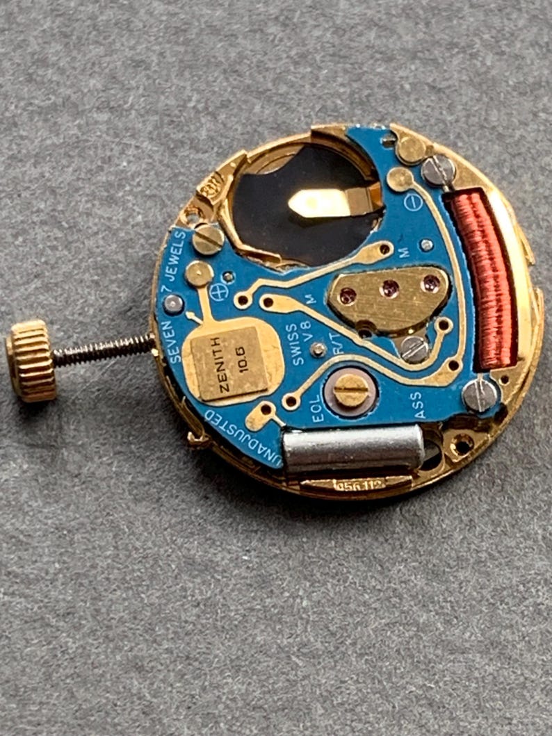 May include: Close-up of a Zenith watch movement. The watch mechanism features a gold-toned frame with a blue circuit board. The text "ZENITH 10.6" and "SWISS V8" are visible. The watch movement also has a gold-toned screw and a red coil.