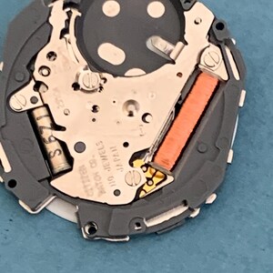 May include: Close-up of a watch movement with intricate mechanical components. The watch mechanism features a gray frame, silver-colored gears, and an orange coil. The text "CITIZEN" and "110 JEWELS JAPAN" is visible.