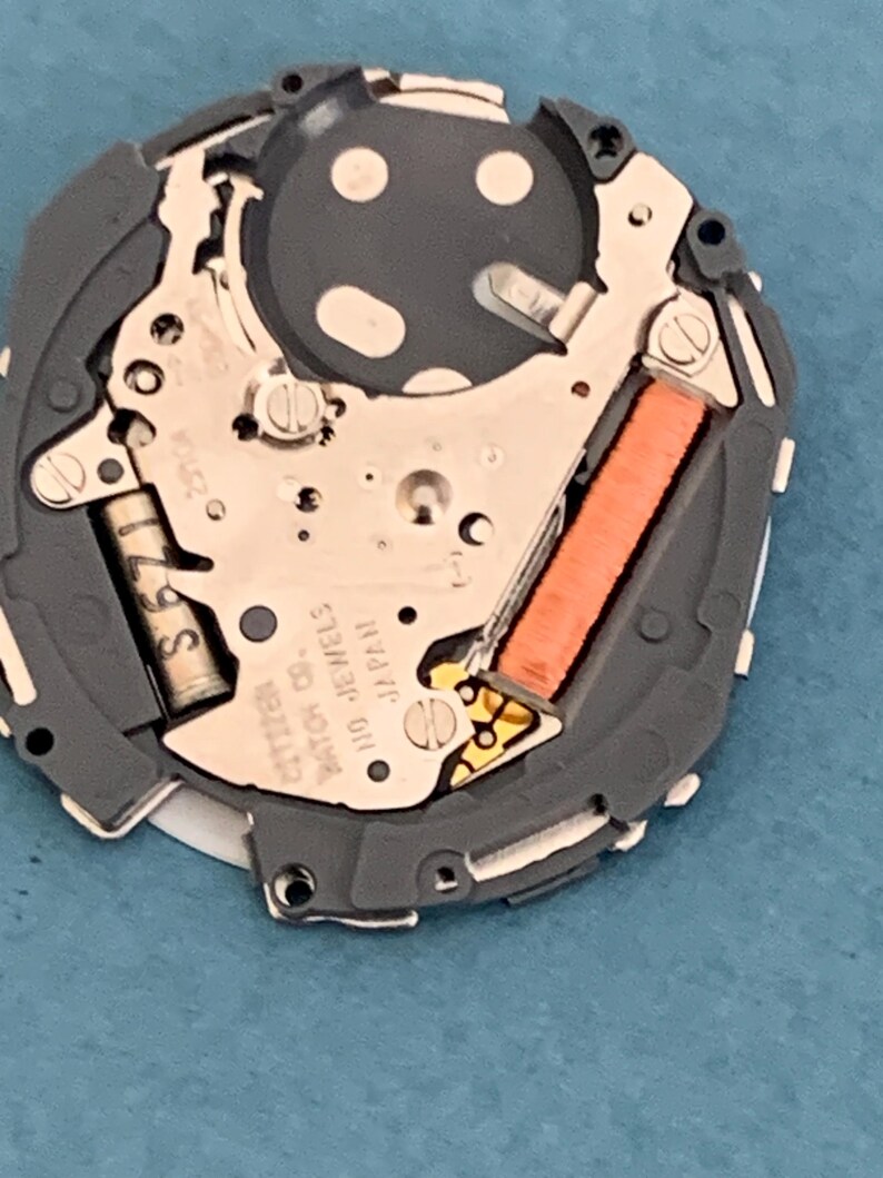 May include: Close-up of a watch movement with intricate mechanical components. The watch mechanism features a gray frame, silver-colored gears, and an orange coil. The text "CITIZEN" and "110 JEWELS JAPAN" is visible.