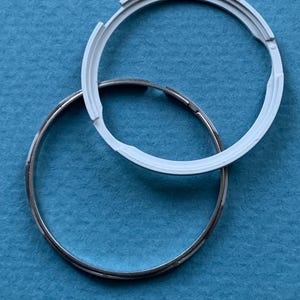 May include: Two circular components on a blue surface. One is a silver metal ring, and the other is a white plastic ring with notched edges. The white ring partially overlaps the silver ring. These appear to be parts of a larger item.