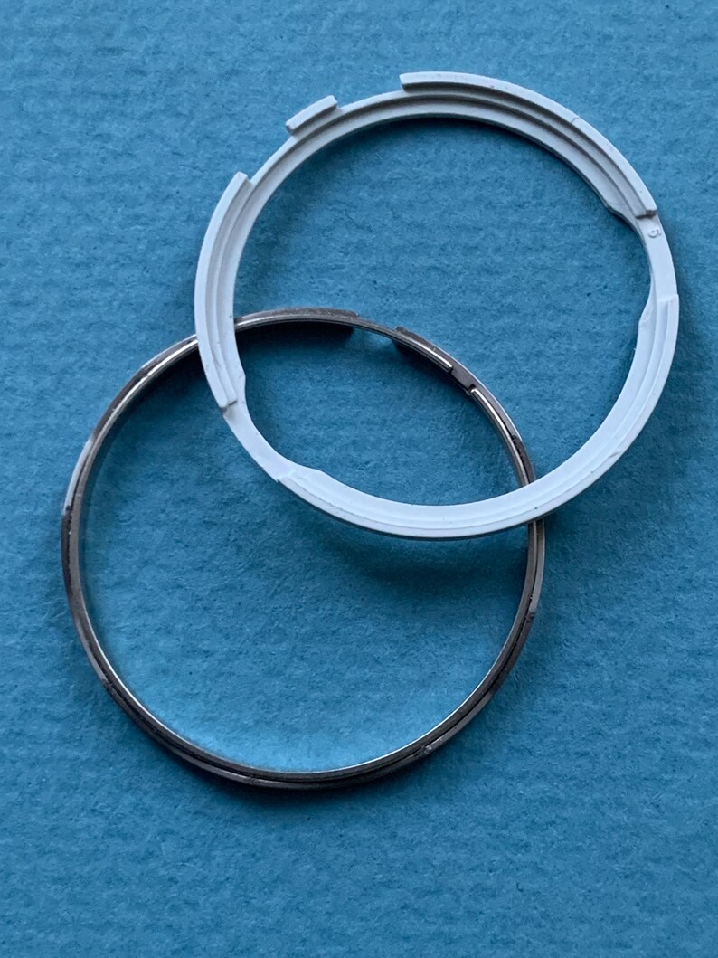May include: Two circular components on a blue surface. One is a silver metal ring, and the other is a white plastic ring with notched edges. The white ring partially overlaps the silver ring. These appear to be parts of a larger item.