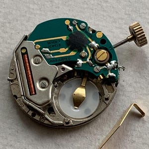 Puede incluir: Primer plano de un movimiento de reloj desmontado. La placa de circuito verde tiene letras doradas que dicen "SEVEN JEWELS" y "UNADJUSTED". Tambi&eacute;n se ven una corona dorada y una peque&ntilde;a palanca dorada.