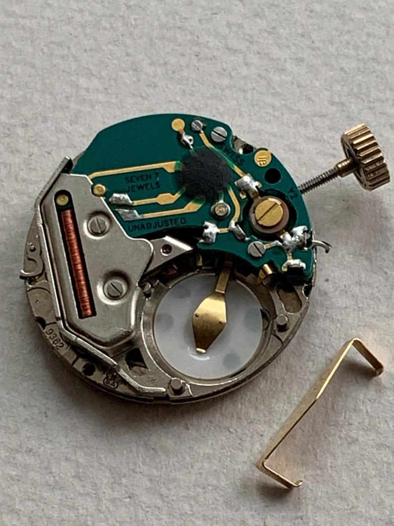 Puede incluir: Primer plano de un movimiento de reloj desmontado. La placa de circuito verde tiene letras doradas que dicen "SEVEN JEWELS" y "UNADJUSTED". Tambi&eacute;n se ven una corona dorada y una peque&ntilde;a palanca dorada.