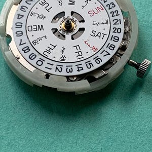 May include: Close-up of a watch movement with a white dial featuring Arabic and English day and date indicators. The dial has a central gear and a silver crown. The background is a teal surface.