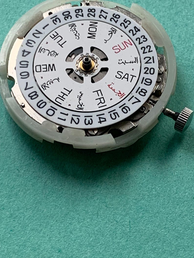 May include: Close-up of a watch movement with a white dial featuring Arabic and English day and date indicators. The dial has a central gear and a silver crown. The background is a teal surface.