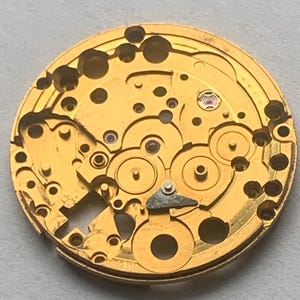 May include: Close-up of a gold-colored watch movement with intricate gears, holes, and small red jewels. The circular mechanism has a complex design, showcasing the internal workings of a timepiece. The watch movement is a vintage item.