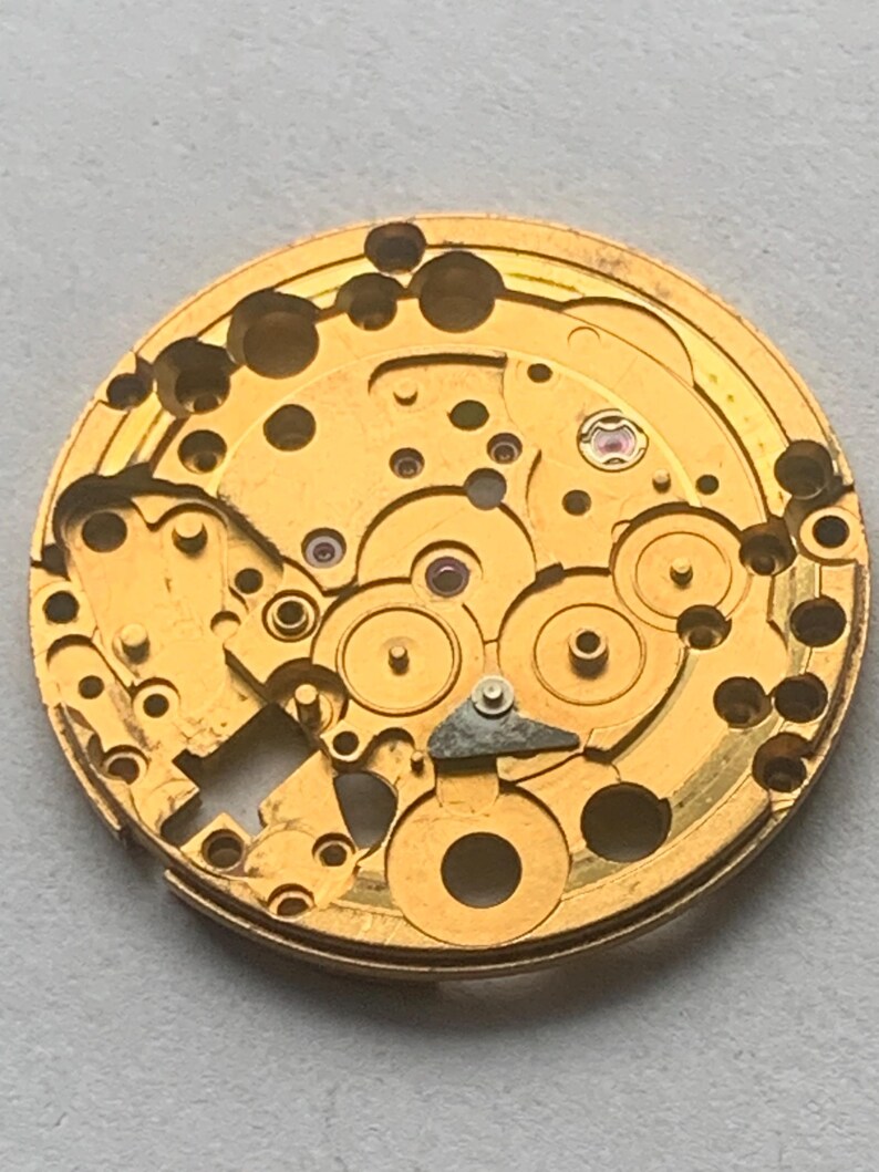 May include: Close-up of a gold-colored watch movement with intricate gears, holes, and small red jewels. The circular mechanism has a complex design, showcasing the internal workings of a timepiece. The watch movement is a vintage item.
