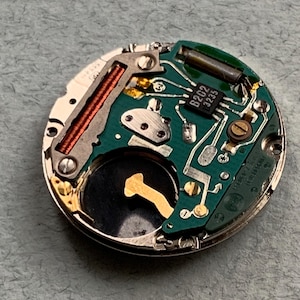 May include: Close-up of a watch movement with a green circuit board, copper coil, and a small black component labeled "B202 3235". The watch mechanism is circular and has various metal components.